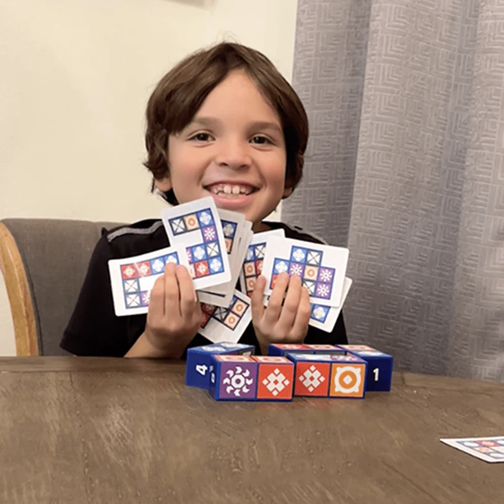 A child smiling and holding a set of patterned cards while sitting at a table.