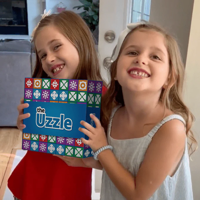 Two smiling children holding the Uzzle game box in a living room.
