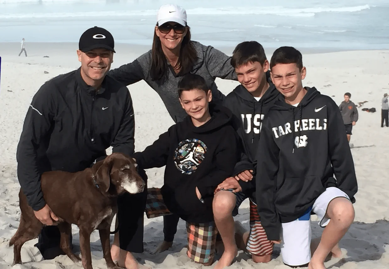 A family with three boys and a dog posing on a sandy beach.
