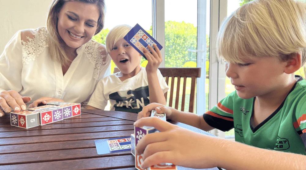 A woman and two children playing a card and dice game at a wooden table.